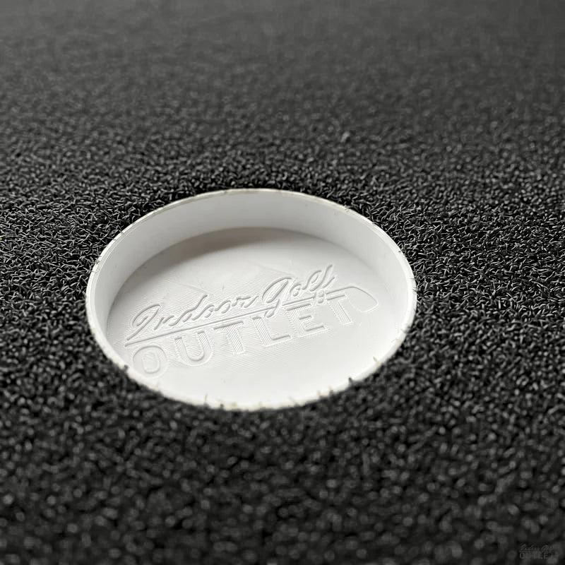 IGO Single-Level Golf Simulator Flooring putting cup sunk in the black turf, embossed with the Indoor Golf Outlet logo.