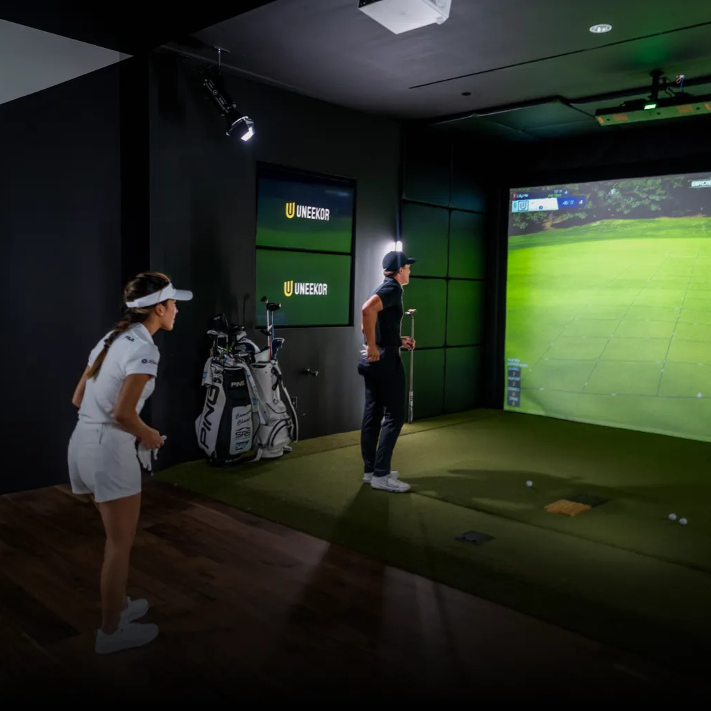 Two people in a Uneekor golf simulation room with an impact screen displaying a golf course.